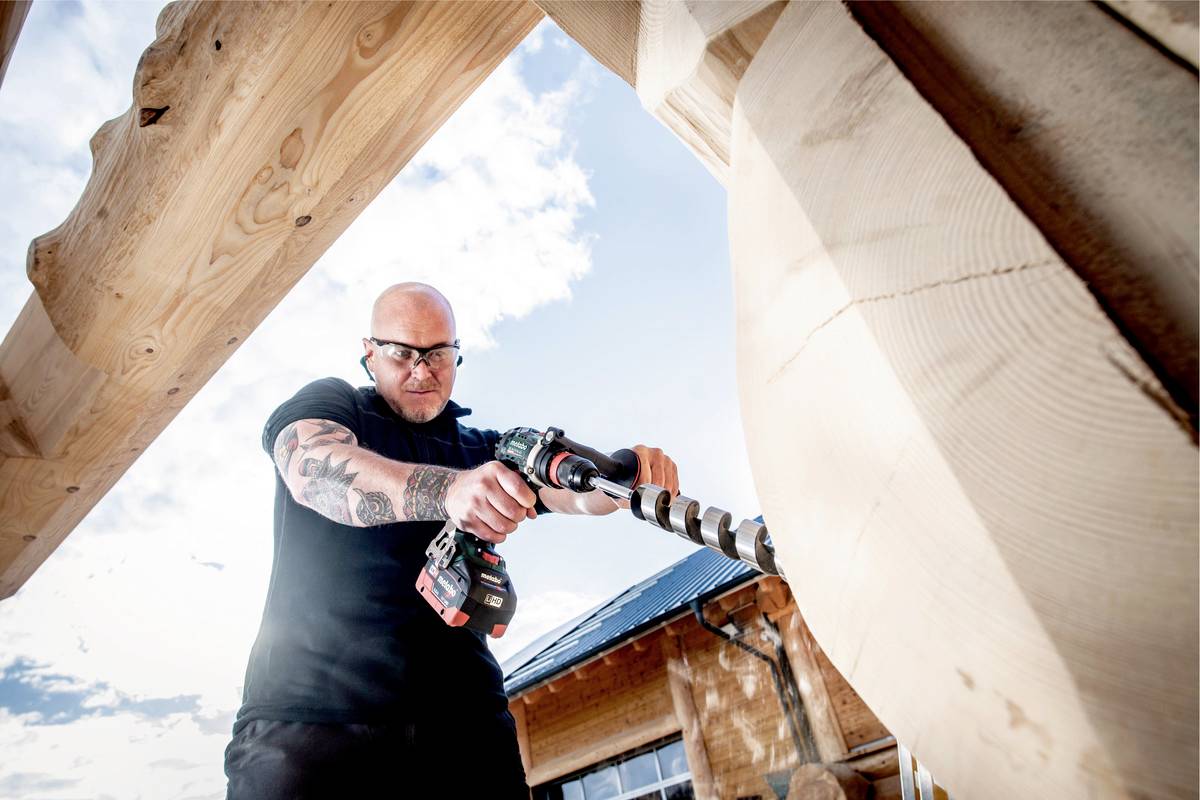 Un homme chauve fore un trou dans un cadre en bois à l'aide d'une grande perceuse. À l'arrière-plan, on peut voir une maison et un ciel bleu.