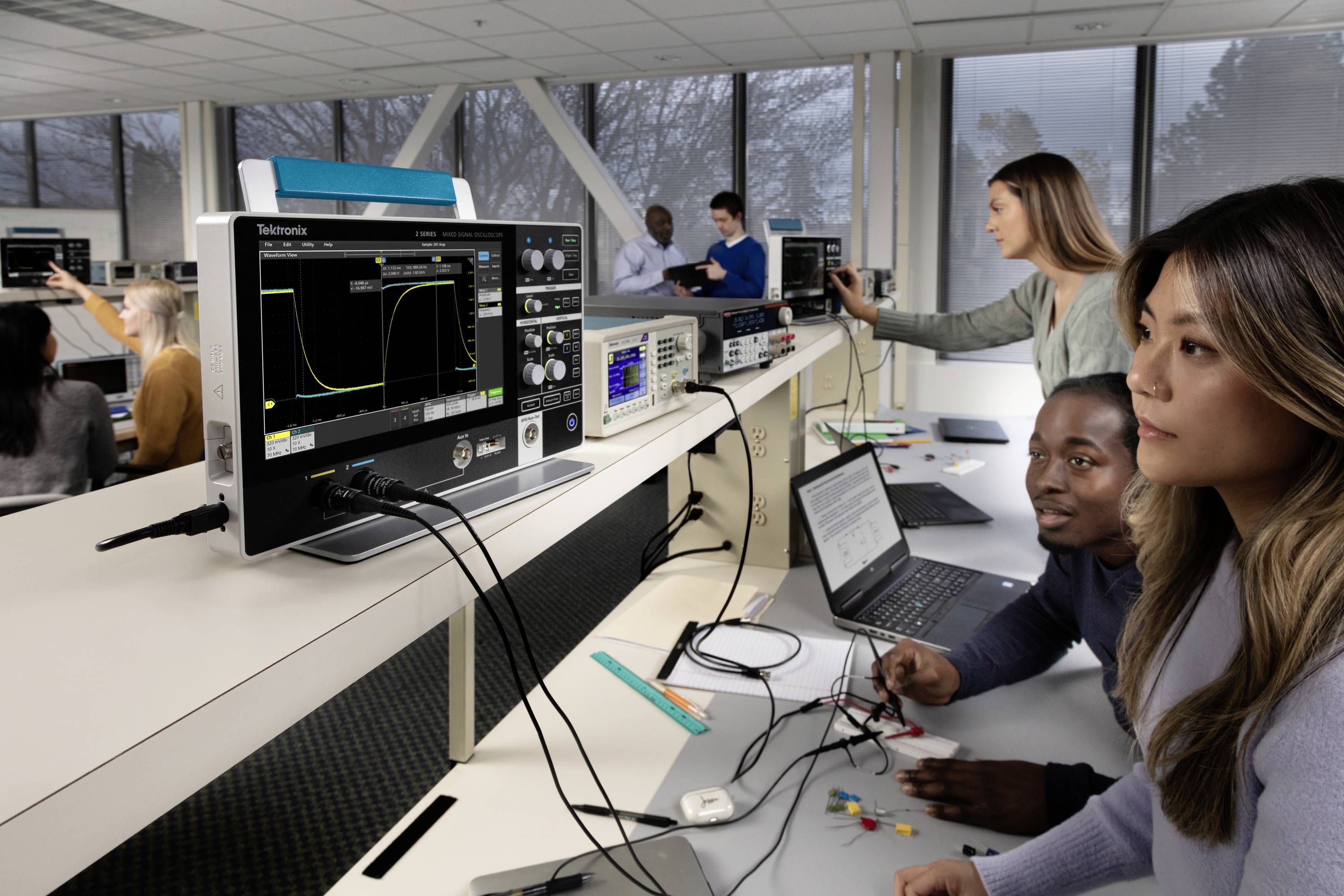 Des personnes travaillent dans un laboratoire d'électronique, utilisant des oscilloscopes sur une table. Les écrans affichent des données de circuits électroniques.