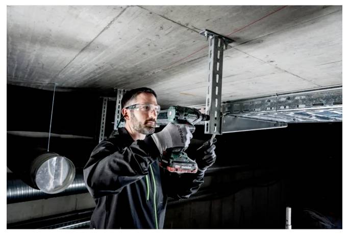 Une personne portant des lunettes de sécurité et des gants utilise une perceuse électrique pour installer des supports de plafond dans un environnement industriel.