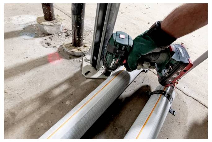 Une personne portant des gants utilise un outil électrique pour fixer des tuyaux métalliques sur un sol. Les tuyaux présentent des lignes jaunes et sont attachés ensemble.