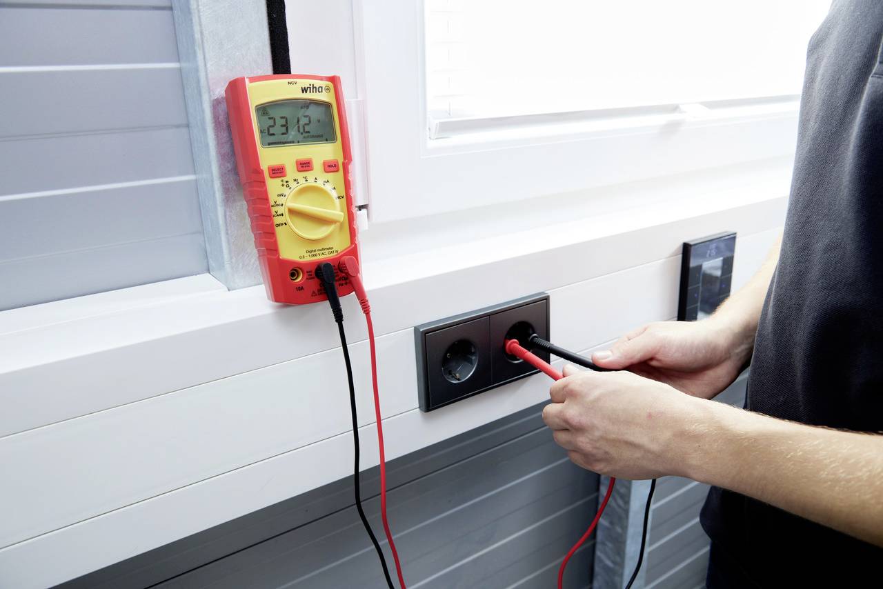 Une personne mesure la tension d'une prise électrique à l'aide d'un multimètre. L'affichage indique '23,12'. La scène se déroule près d'un encadrement de fenêtre.