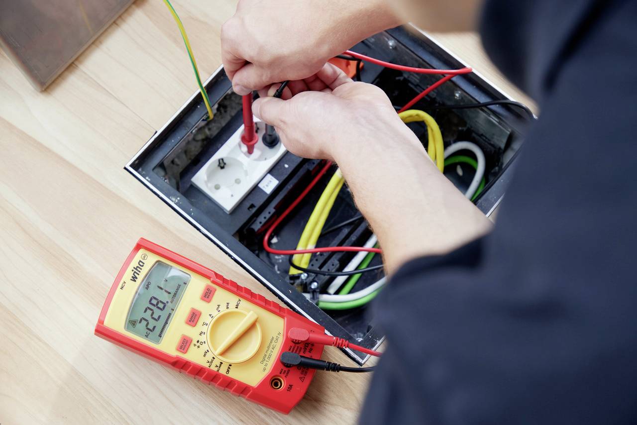 Une personne utilise un multimètre pour vérifier les connexions électriques dans une prise de courant. Plusieurs câbles colorés sont visibles.