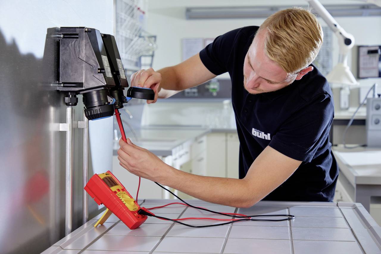 Un homme travaille dans un laboratoire, testant des connexions électriques à l'aide d'un multimètre. Il mesure la résistance d'un appareil.