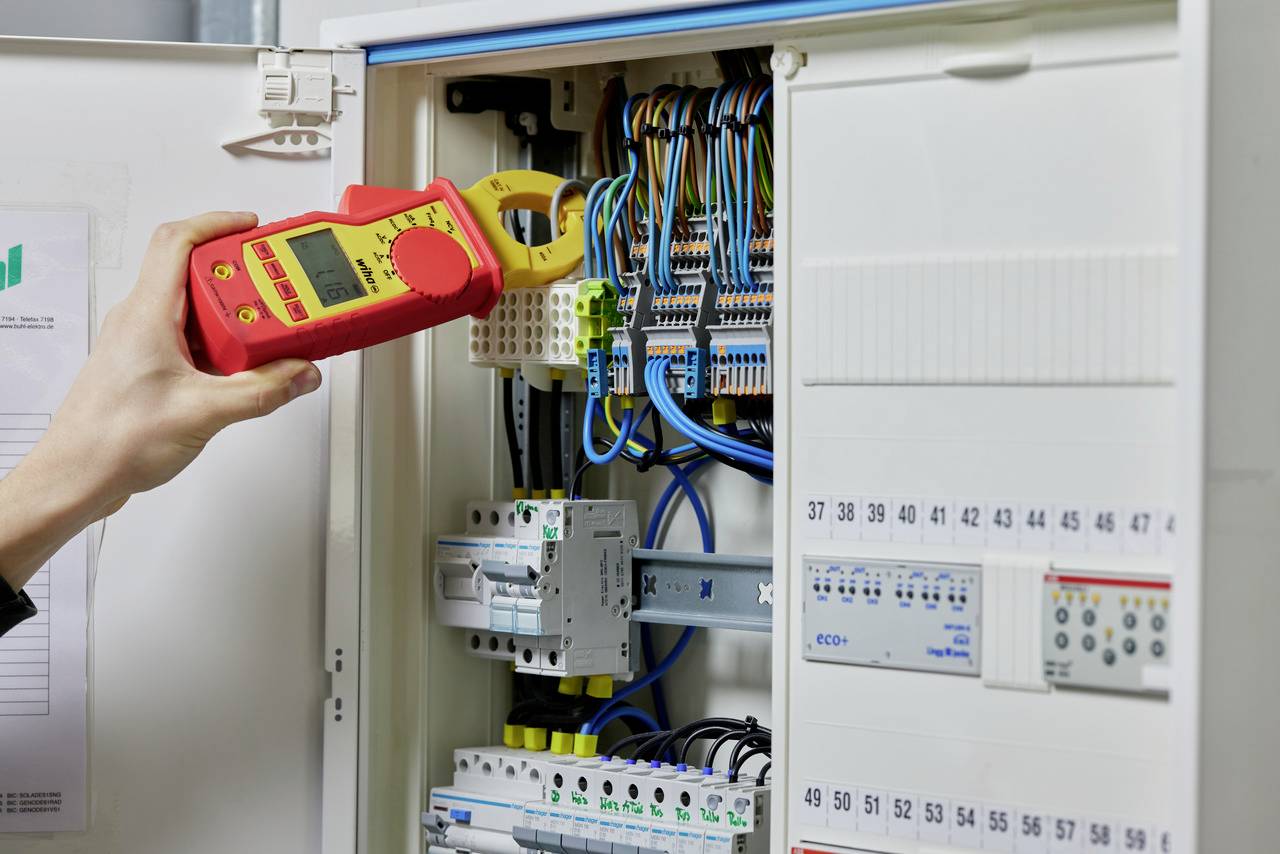 Une personne mesure la tension électrique dans un armoire électrique ouverte à l'aide d'un multimètre. De nombreux câbles colorés sont visibles.