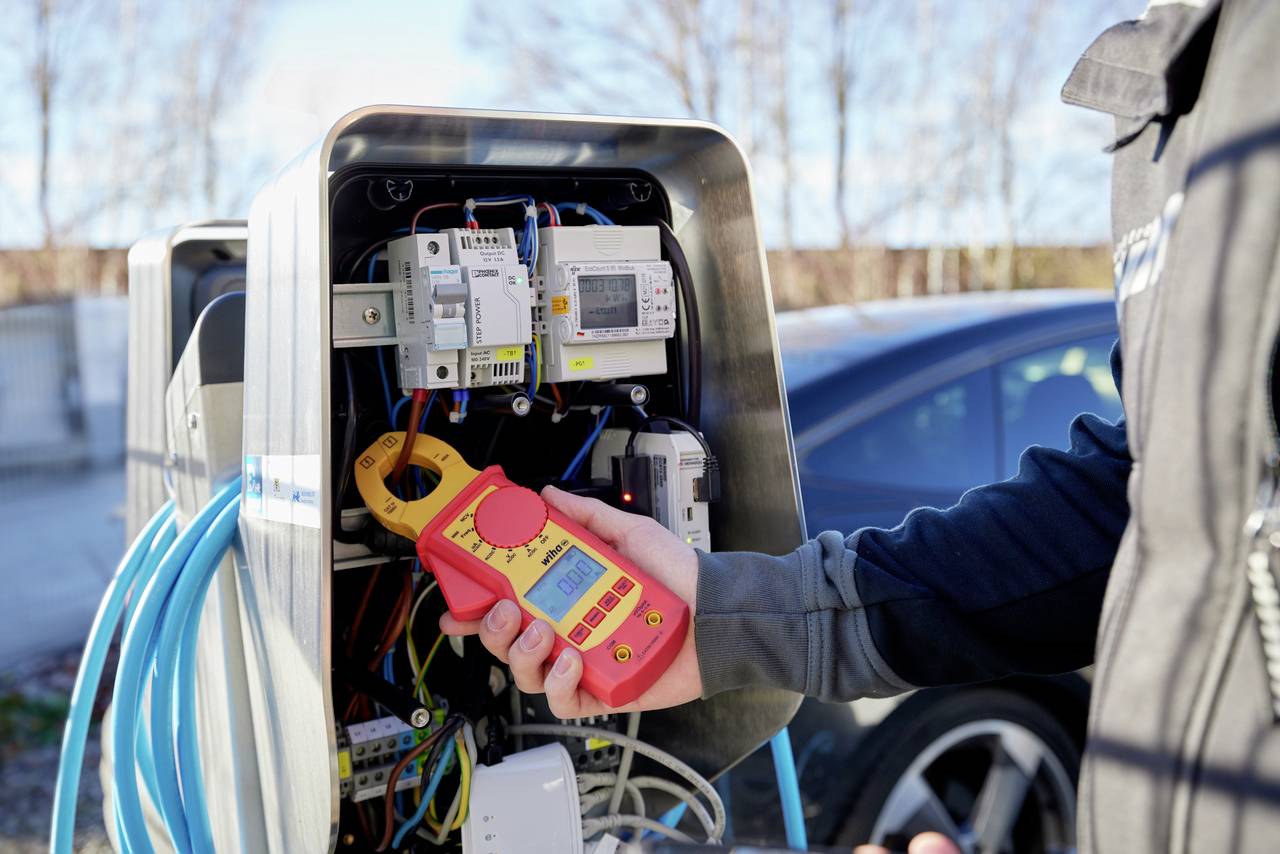 Une personne tient un appareil de mesure à côté d'une borne de recharge pour véhicules électriques ouverte et vérifie la connexion électrique.
