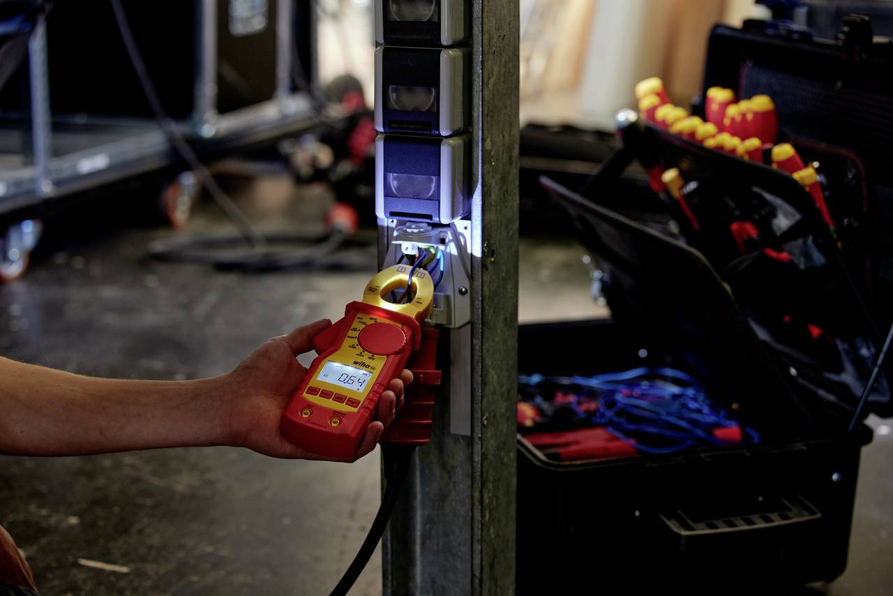 Une personne mesure le courant électrique dans un câble à l'aide d'un multimètre numérique. Un coffret à outils contenant des tournevis est visible en arrière-plan.