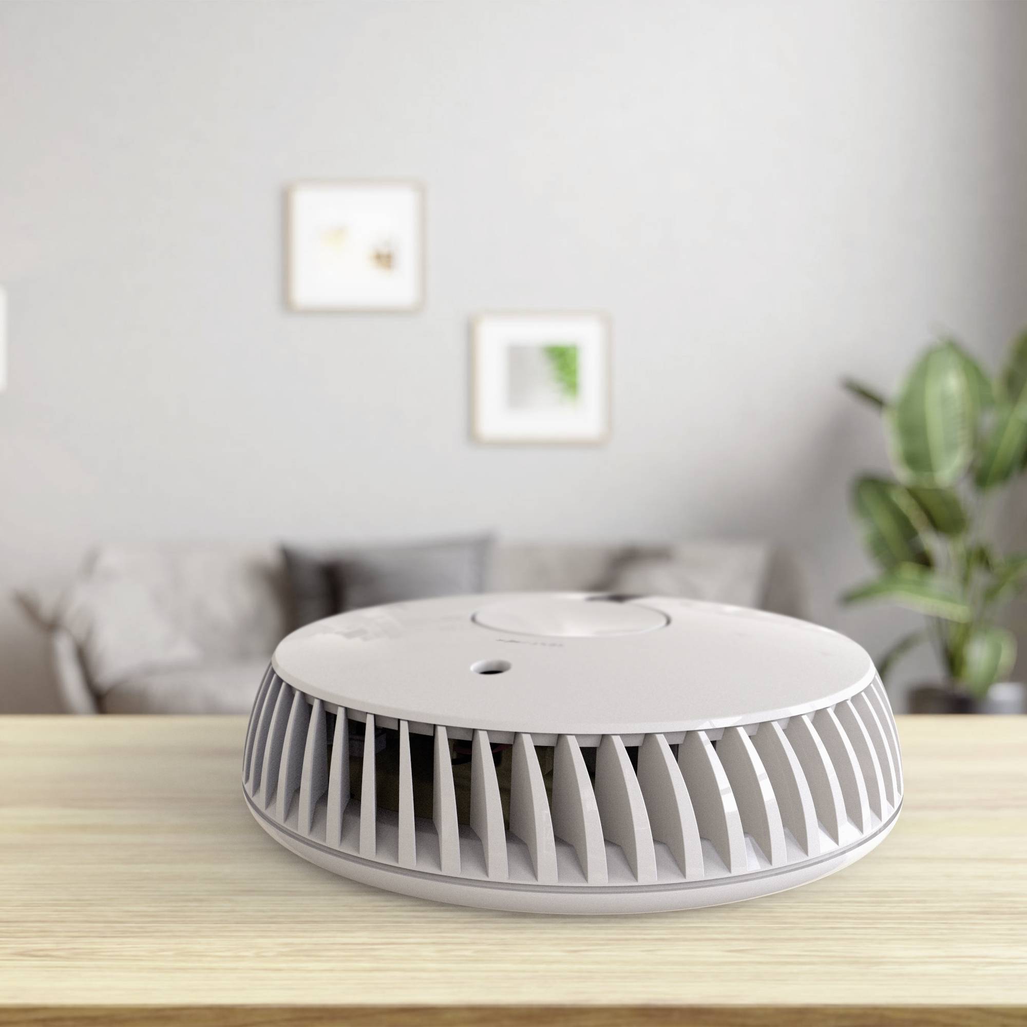 Un petit purificateur d'air rond est posé sur une table en bois. En arrière-plan, on peut voir un canapé, des plantes et des tableaux accrochés au mur.