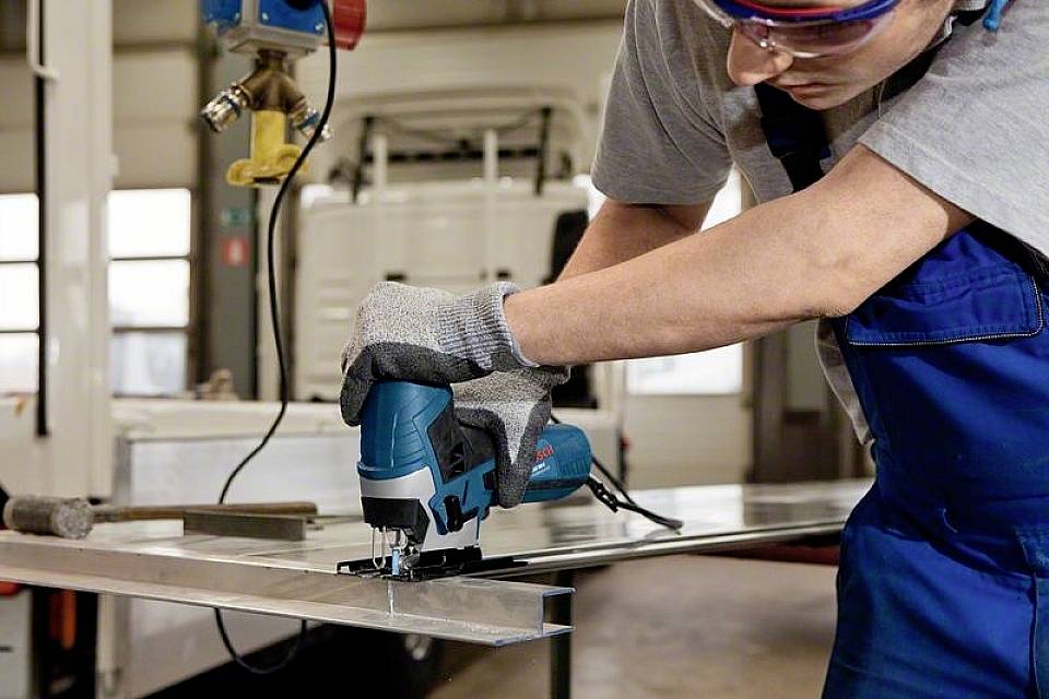 Un ouvrier coupe une plaque métallique avec une scie sauteuse électrique dans un atelier, portant des lunettes de protection et des gants.