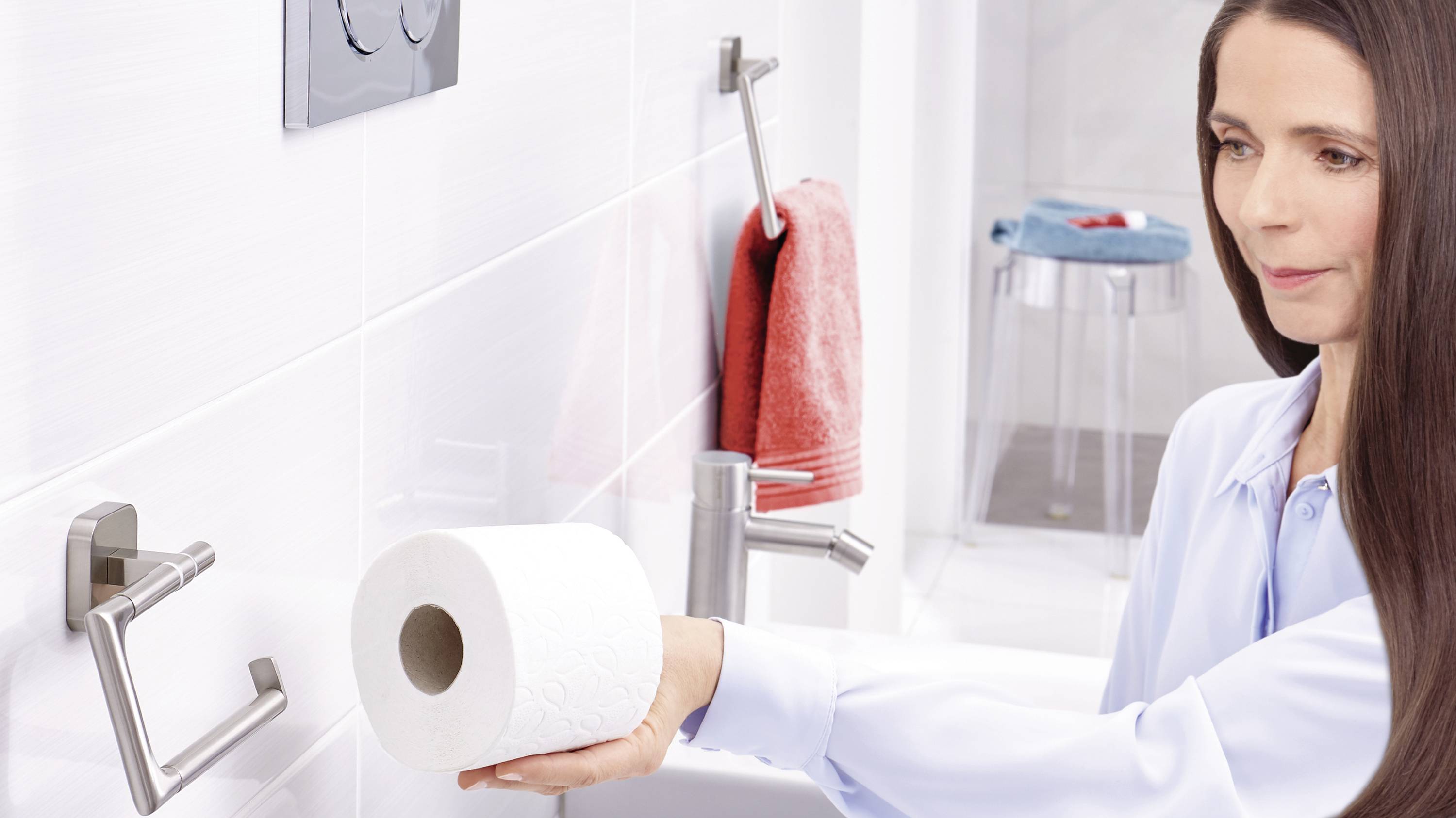 Une femme change un rouleau de papier toilette dans la salle de bain. Elle porte une chemise claire et se tient devant un lavabo avec un porte-serviettes.