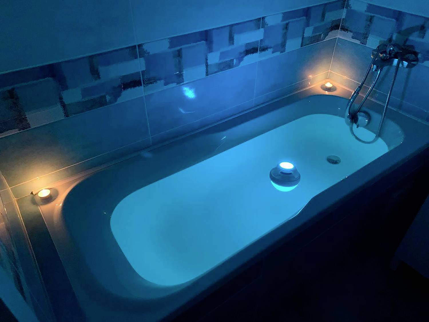 Une baignoire remplie, éclairée par une faible lumière bleue et bordée de bougies, crée une atmosphère relaxante.