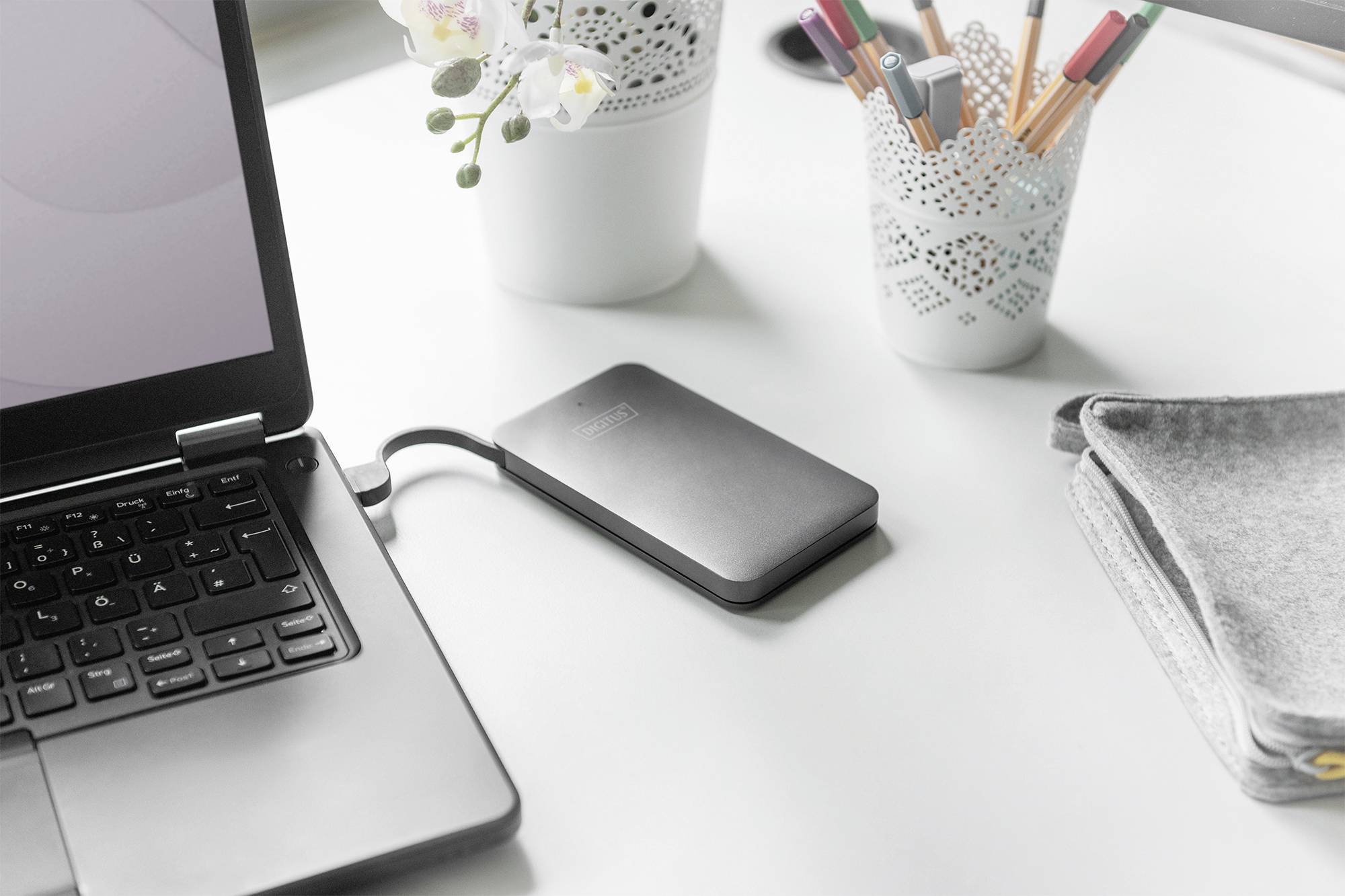 Disque dur externe connecté à un ordinateur portable sur un bureau, avec des stylos et des plantes en arrière-plan.