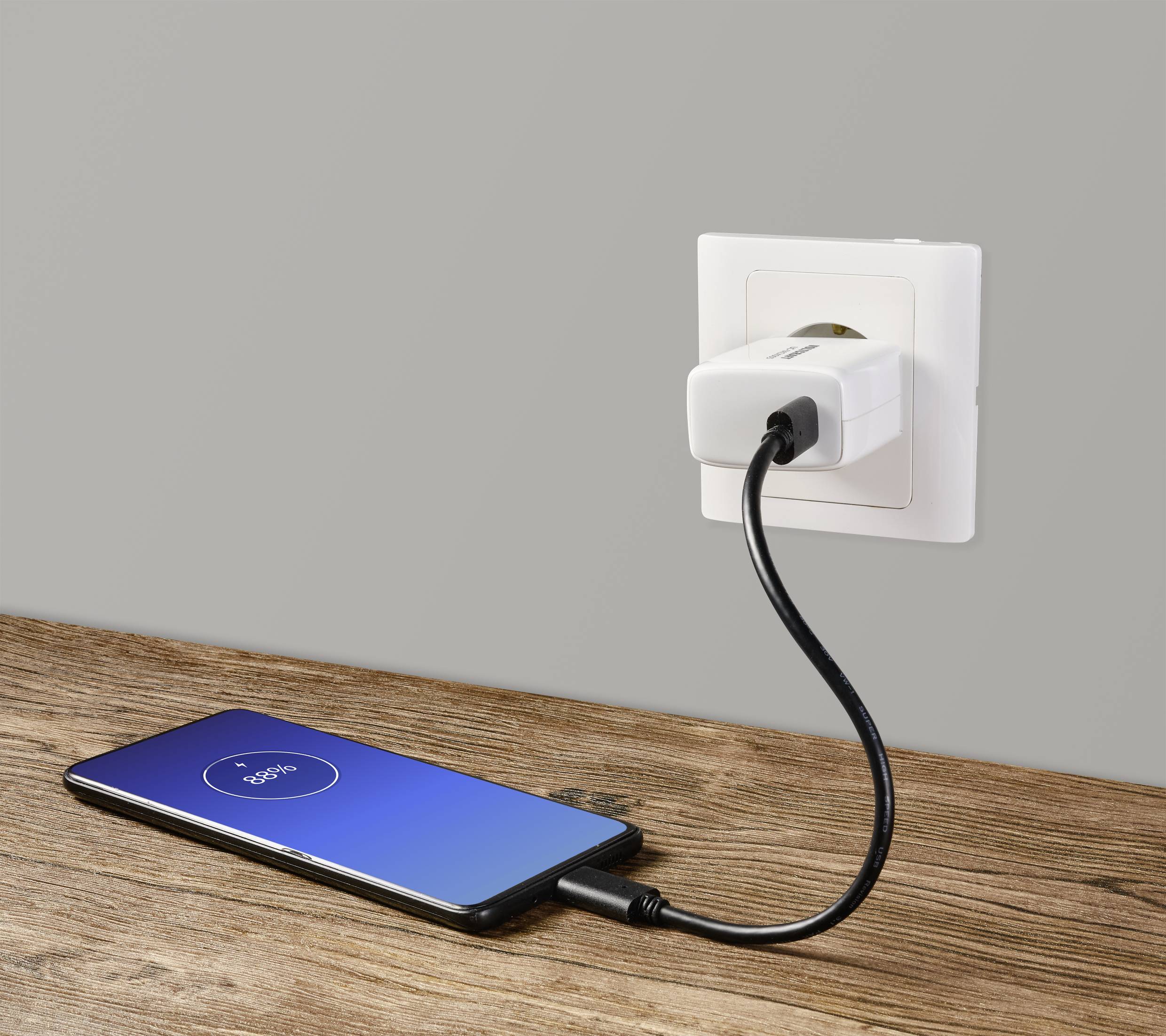 Un smartphone est posé sur une table en bois et se charge, connecté par un câble à une prise électrique blanche sur un mur gris.