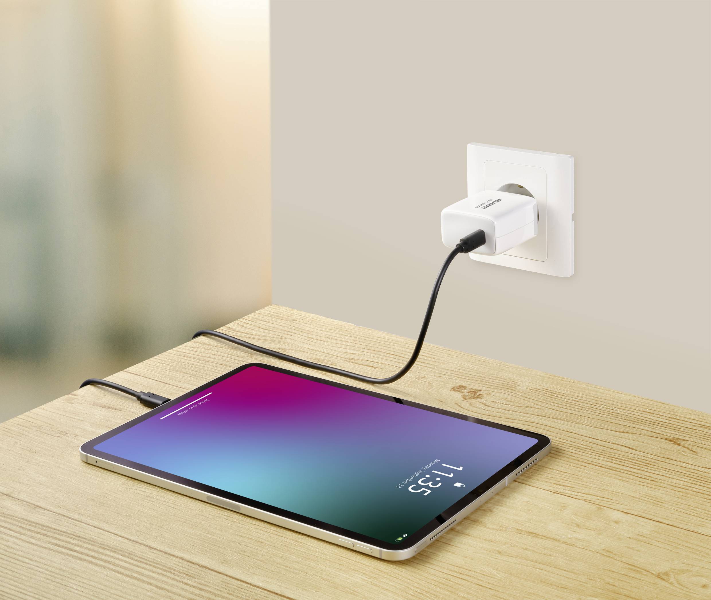 Une tablette est posée sur une table et se charge via un câble branché à une prise électrique. L'écran affiche l'heure 11:35.