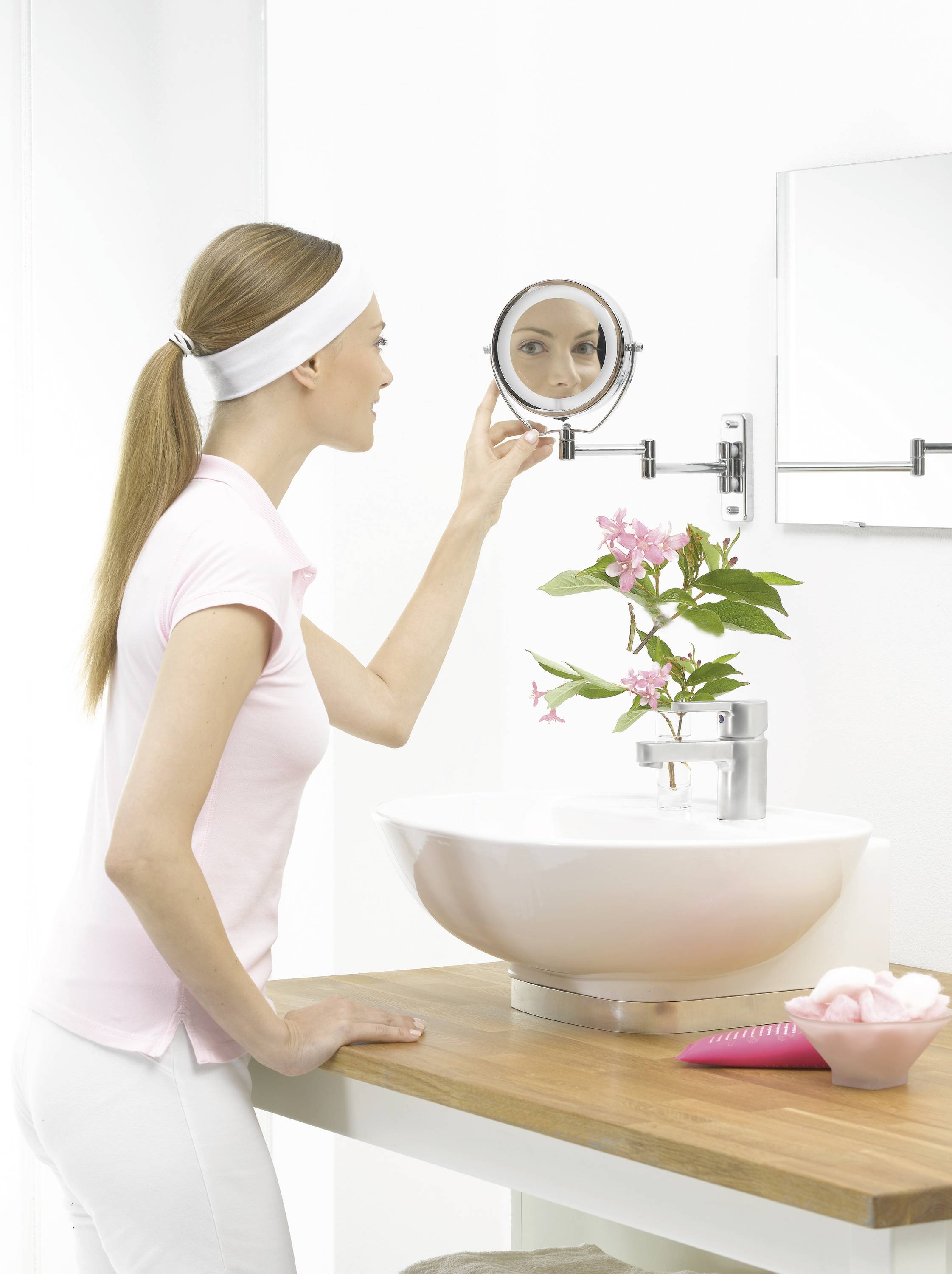 Une femme se regarde dans un miroir mural rond dans la salle de bain, entourée de murs blancs et d'une fleur dans un vase à côté du lavabo.