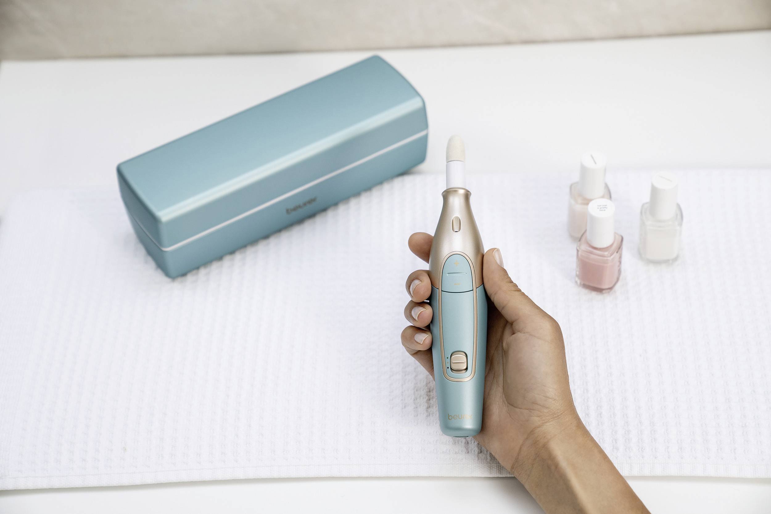 Une main tient un appareil électrique de soin des ongles à côté de ongles vernis et de trois flacons de vernis à ongles, avec un étui en arrière-plan.