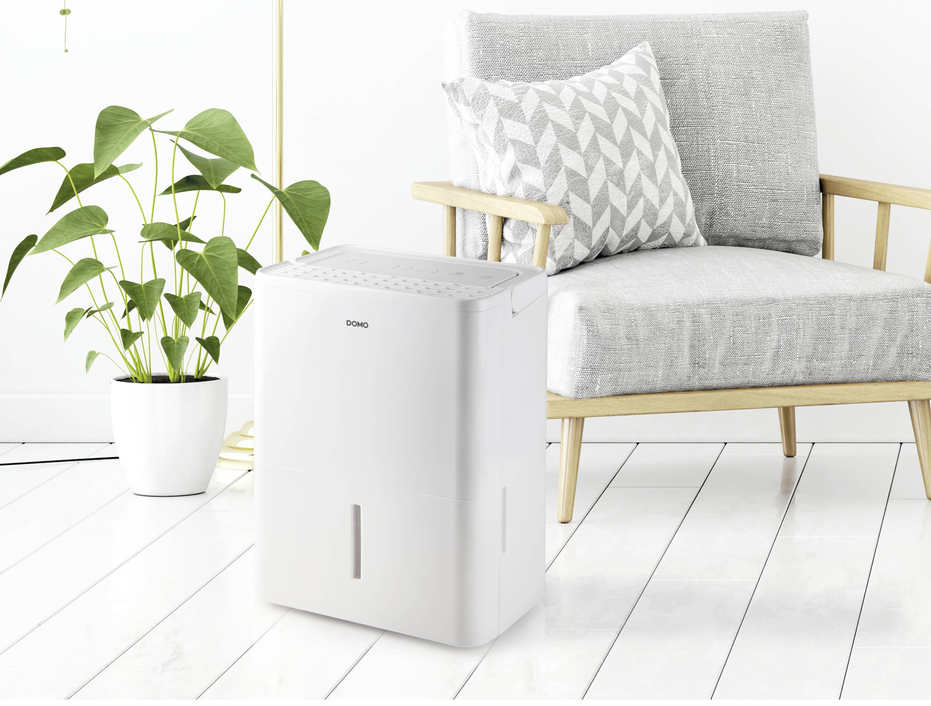 Un déshumidificateur blanc est posé sur un sol en bois clair, à côté d'un fauteuil gris et d'une plante en pot dans une pièce moderne.