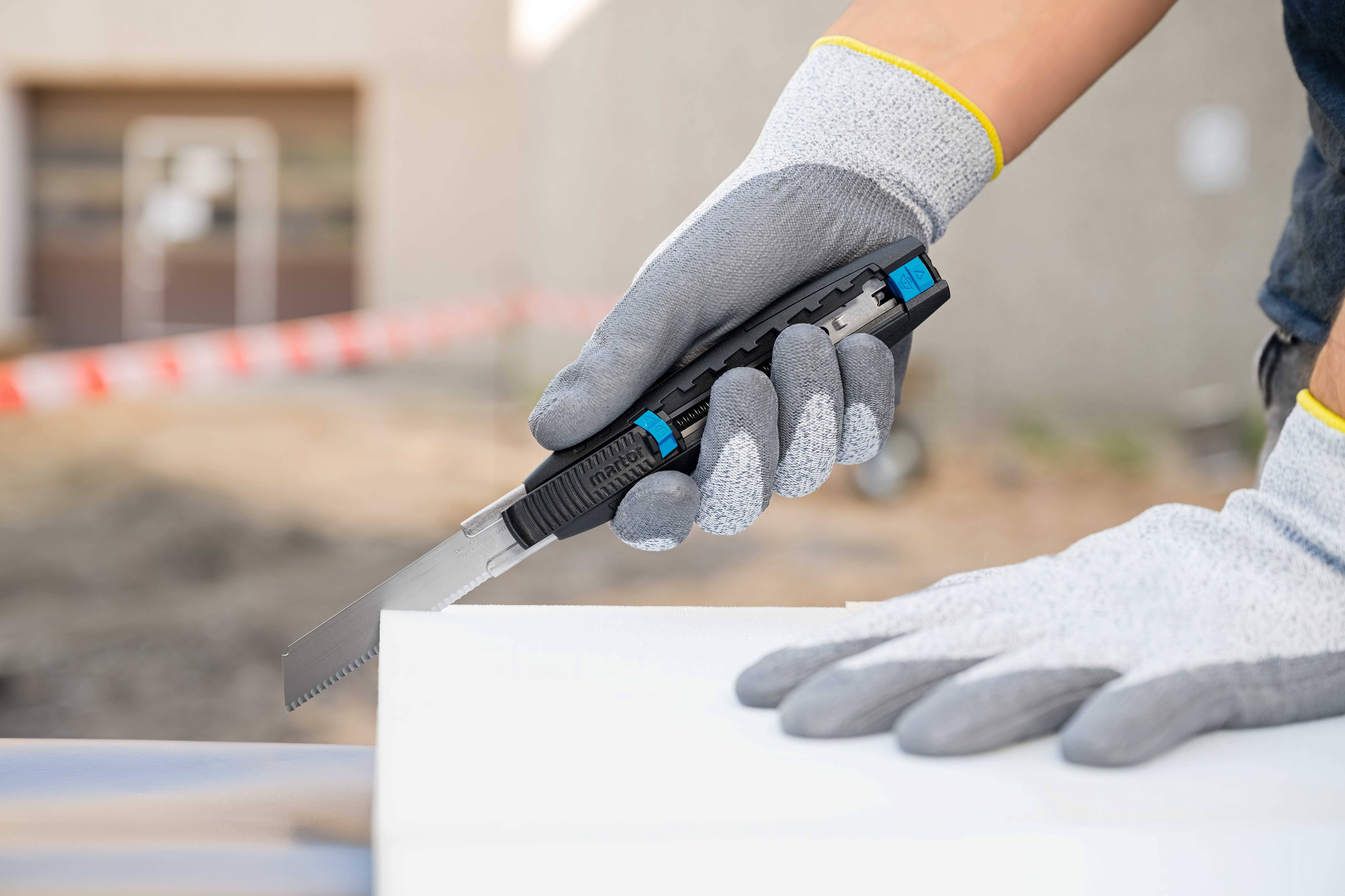 Une personne portant des gants gris utilise un couteau à tapis pour couper un matériau blanc. Arrière-plan flou.