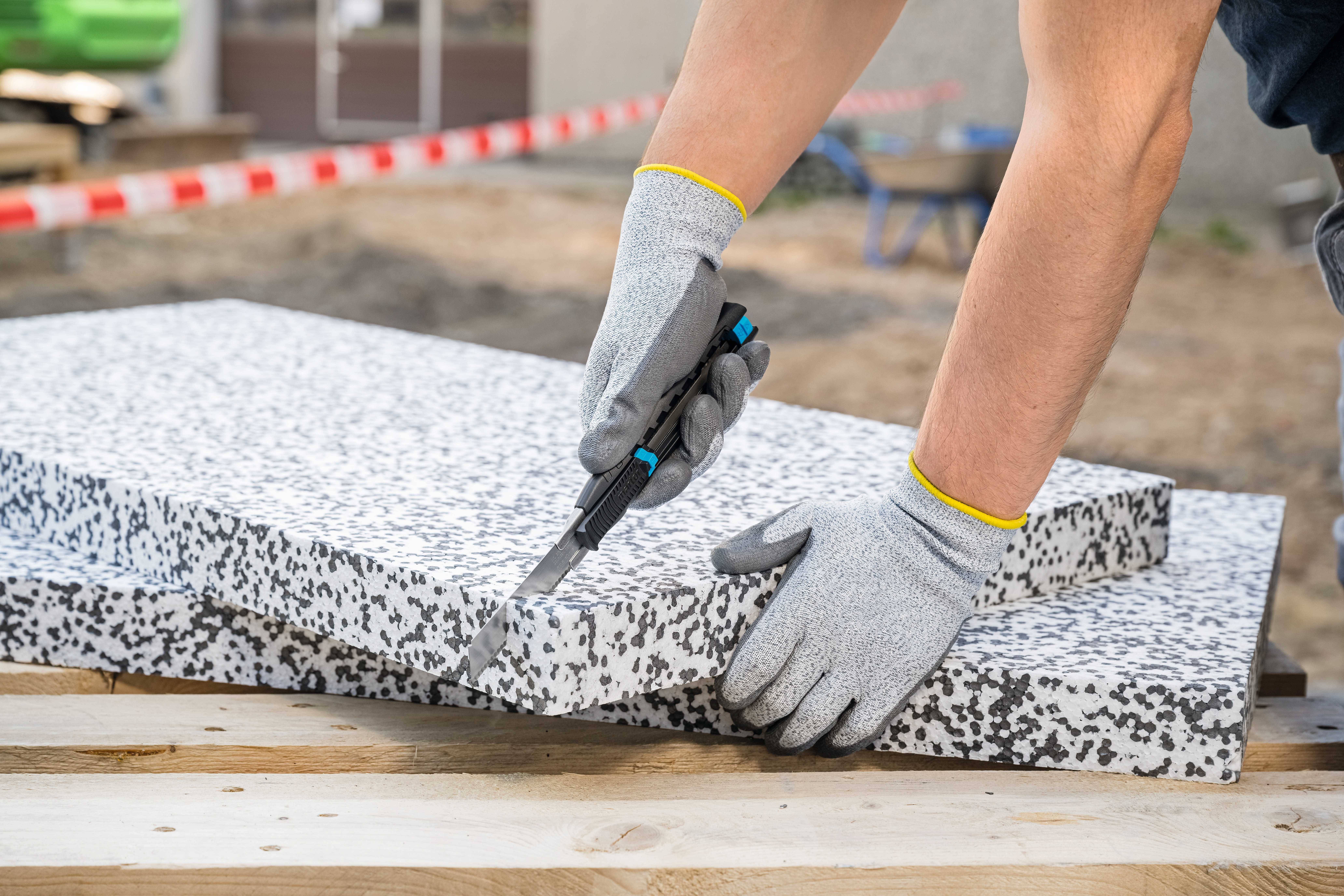 Une personne portant des gants gris travaille le bord d'une dalle de pierre grossièrement texturée à l'aide d'un outil sur un chantier de construction.