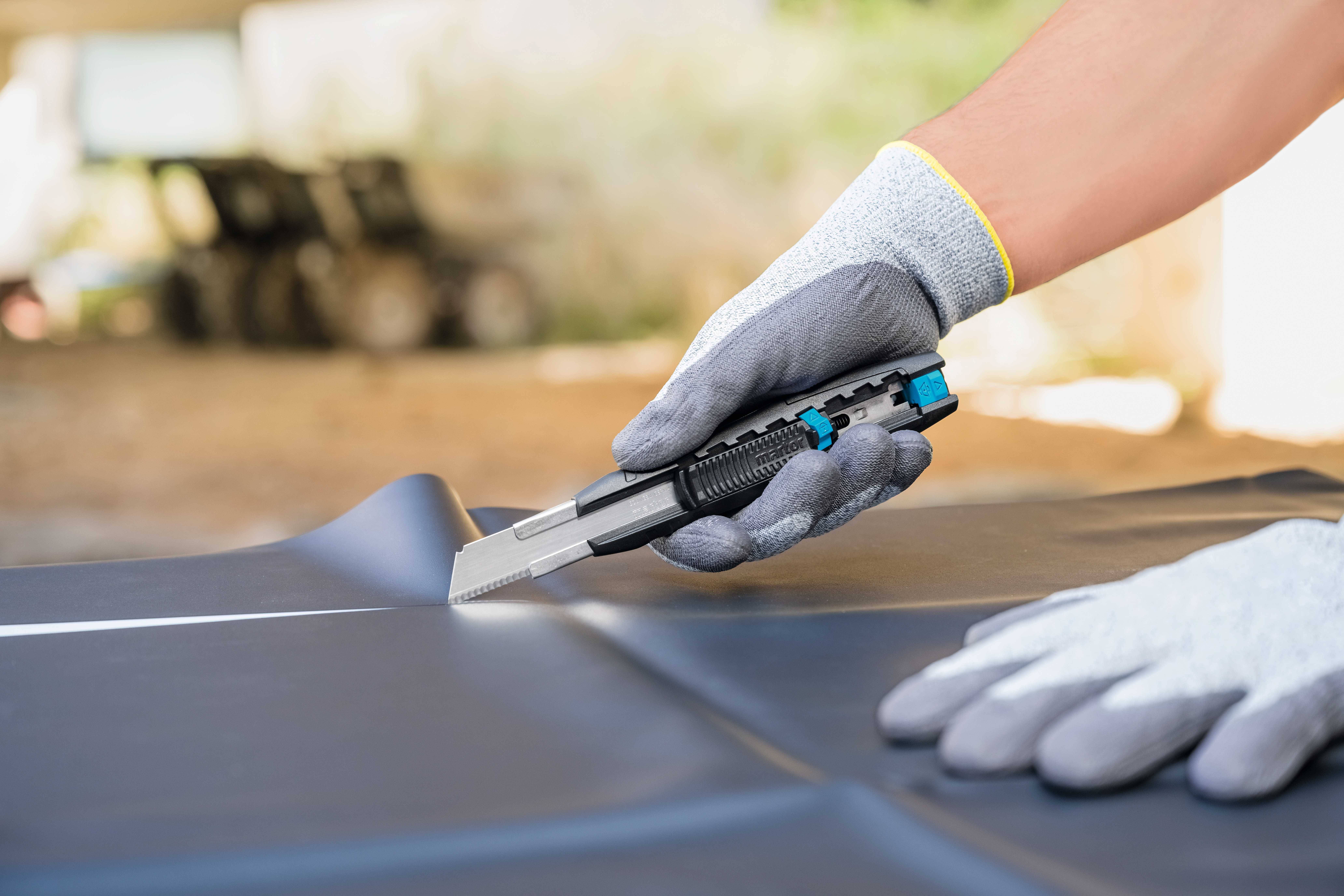 Une personne portant des gants gris coupe un tapis noir avec un cutter sur une table. Arrière-plan flou.