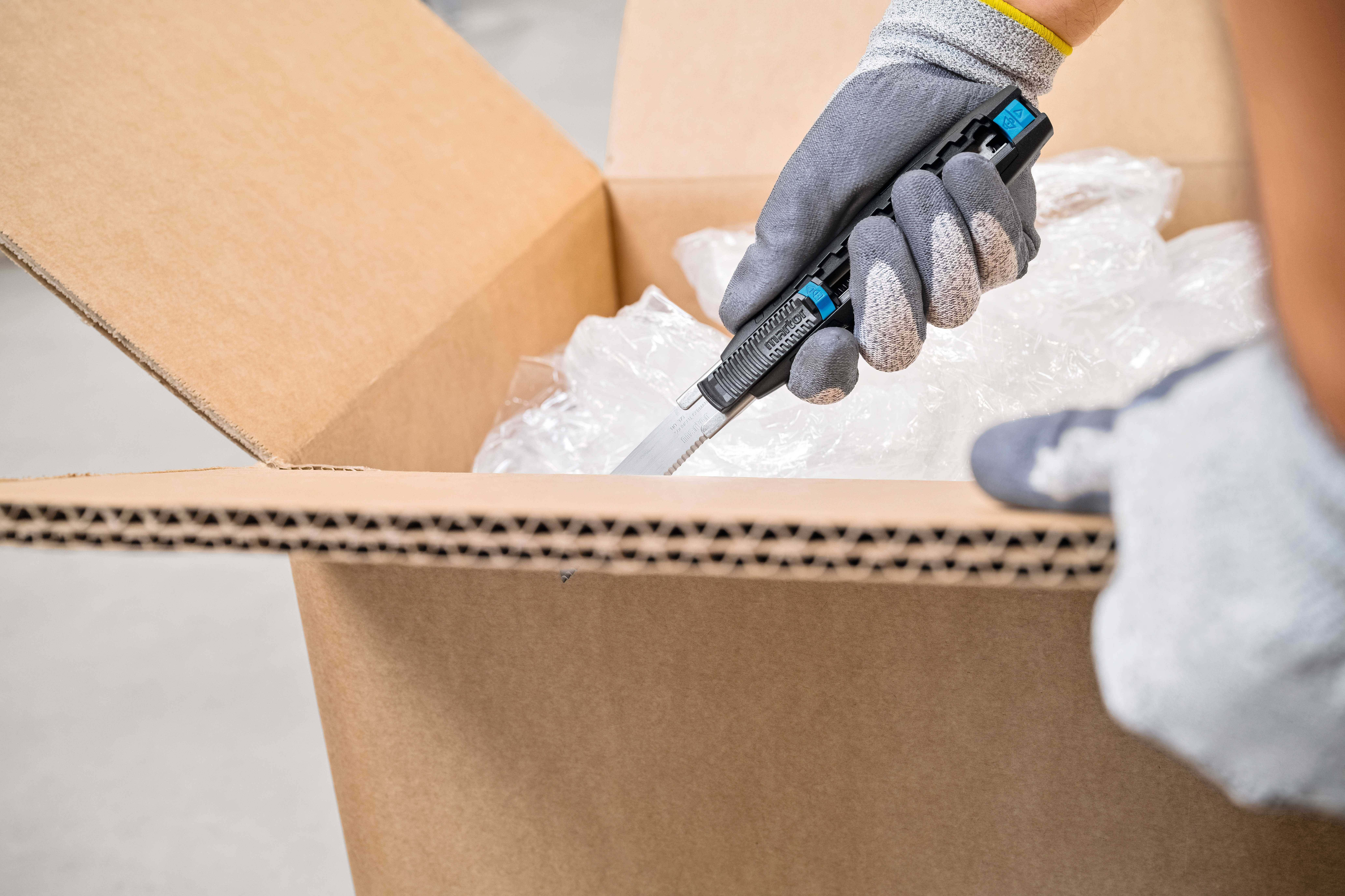 Une personne portant des gants gris ouvre prudemment un carton en carton à l'aide d'un cutter. Le carton contient du film à bulles.