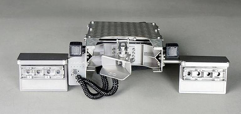 Un appareil argenté, rectangulaire, doté de deux bras escamotables et de câbles, dans un environnement industriel. Il semble s'agir d'un équipement technique.