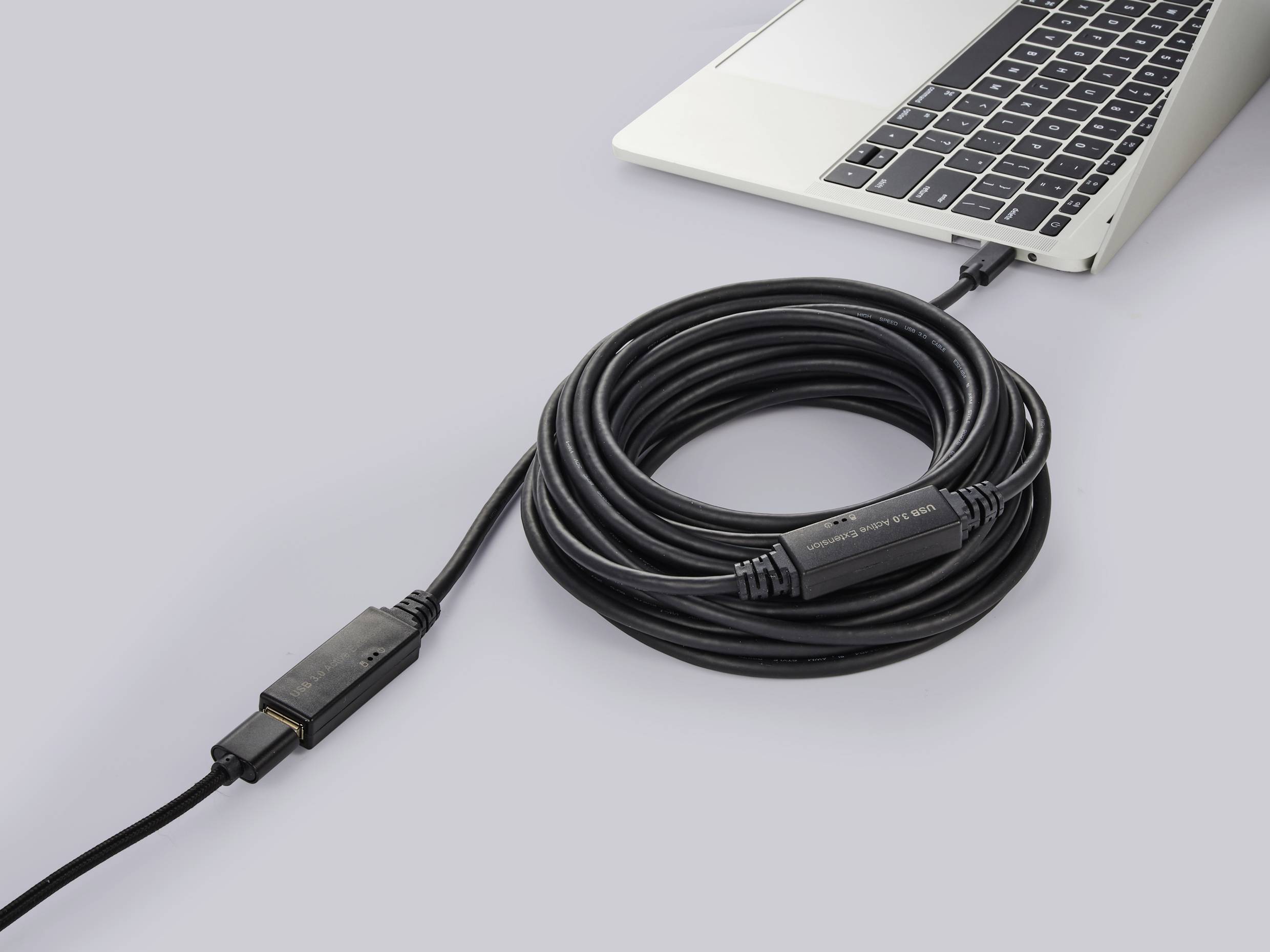 Un câble HDMI noir est connecté à un ordinateur portable ouvert. Le câble est enroulé et équipé d'un amplificateur en son milieu.