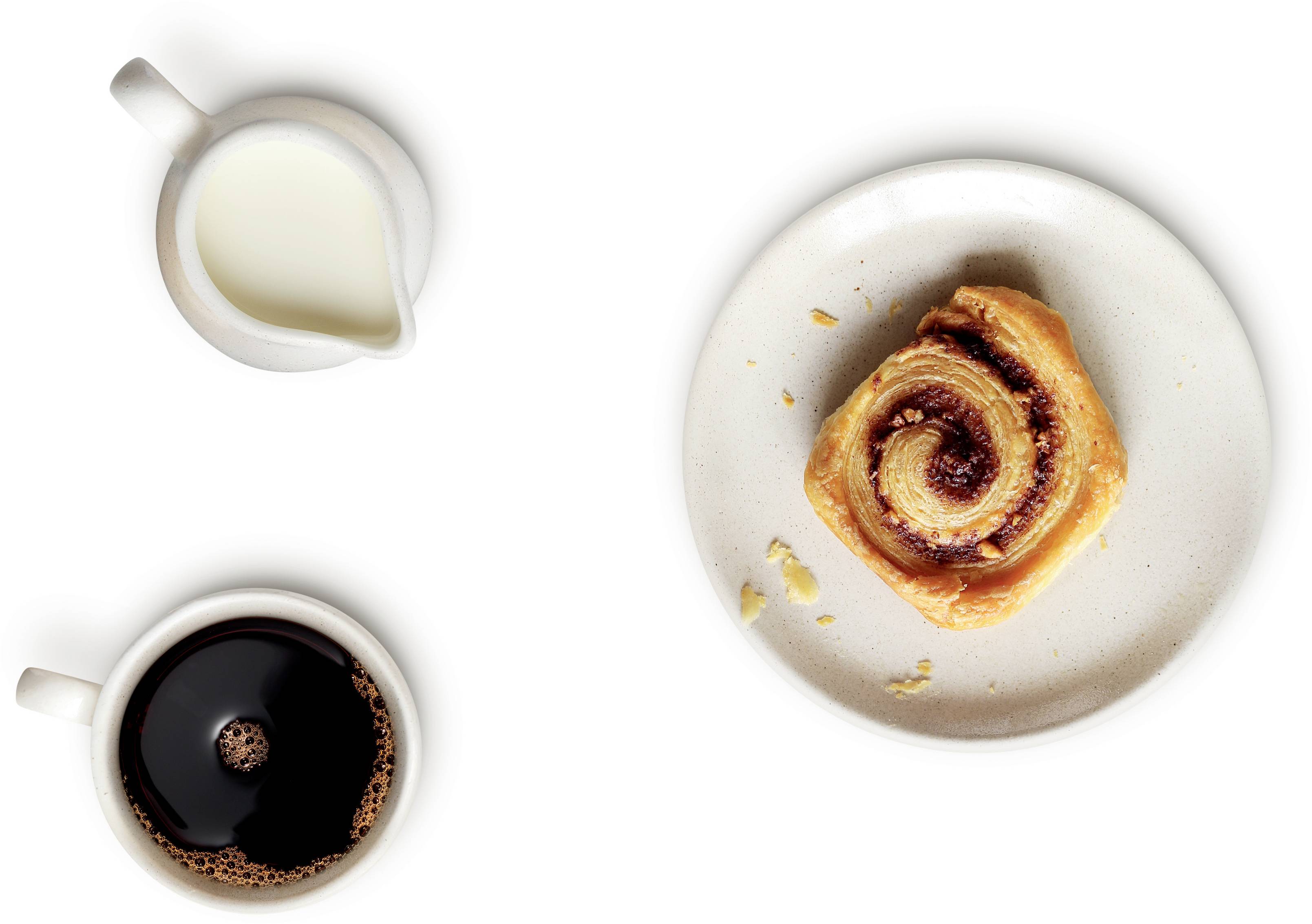 Tasse de café, pot de lait et un pastry roulé à la cannelle sur une assiette.