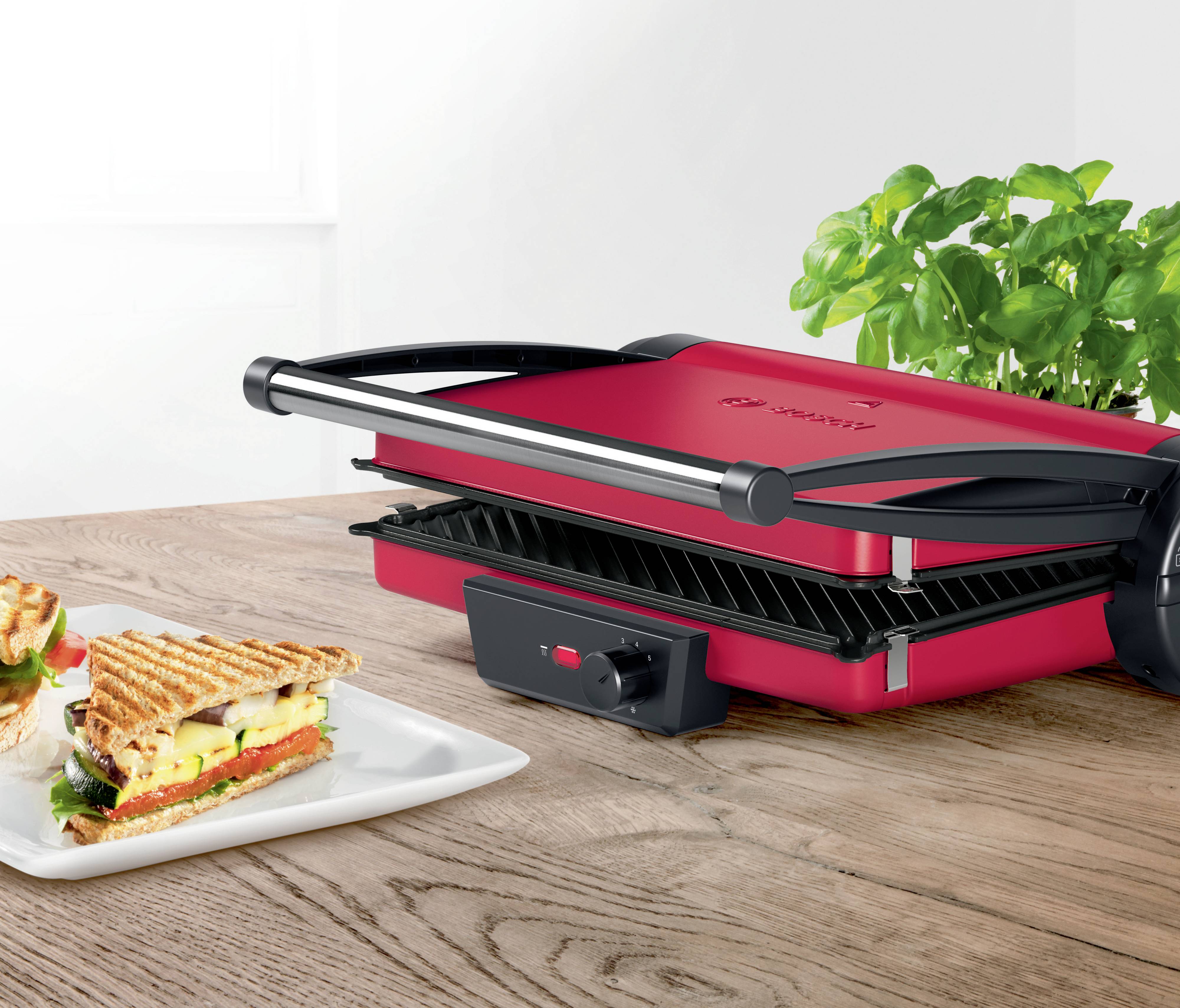 Un grill de contact rouge sur une table en bois à côté de basilic frais, avec un sandwich grillé sur une assiette devant.