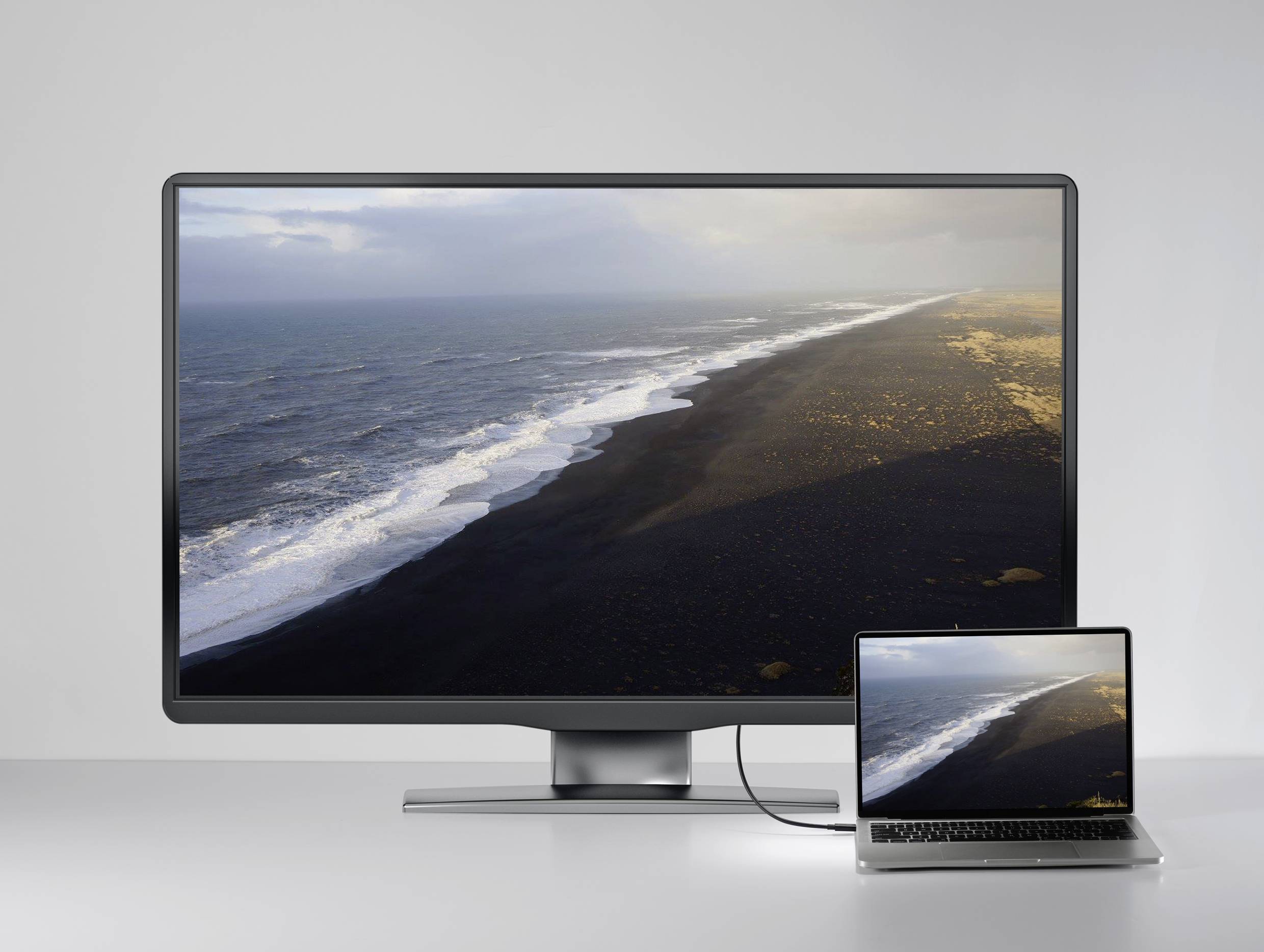 Un grand moniteur et un ordinateur portable connecté affichent la même image d'une ligne de côte avec du sable noir et des vagues.