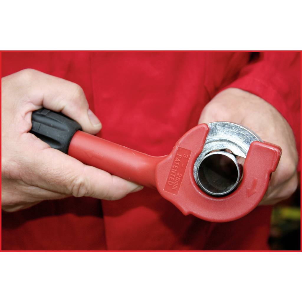 Un outil de coupe de tuyaux rouge tenu dans la main d'une personne est utilisé pour couper un tuyau métallique. Des gants protègent les mains.