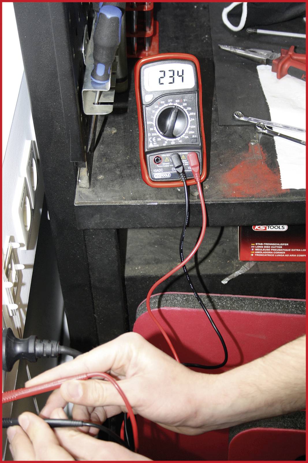 Un multimètre mesure une tension électrique de 234 volts. Au premier plan, une main tient les câbles de mesure sur une connexion. Environnement : zone d'atelier.