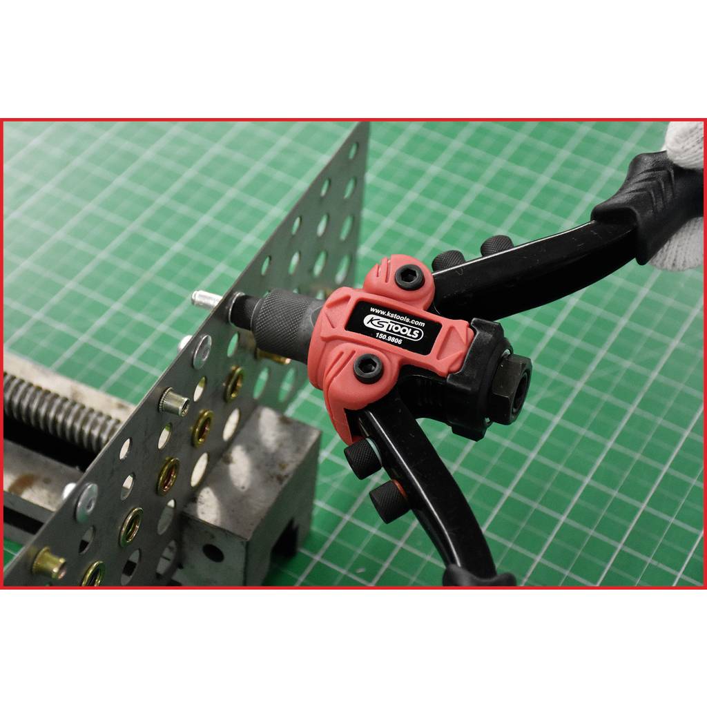 Une pince coupante rouge et noire est utilisée pour travailler une plaque métallique percée de plusieurs trous, sur un fond quadrillé vert.