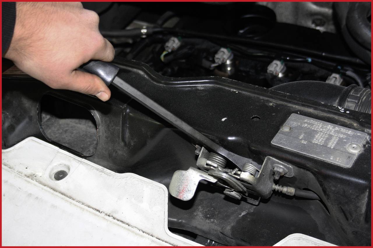Une main tient une tige métallique et tire sur une pièce noire d'automobile dans le compartiment moteur, fixée par des vis.