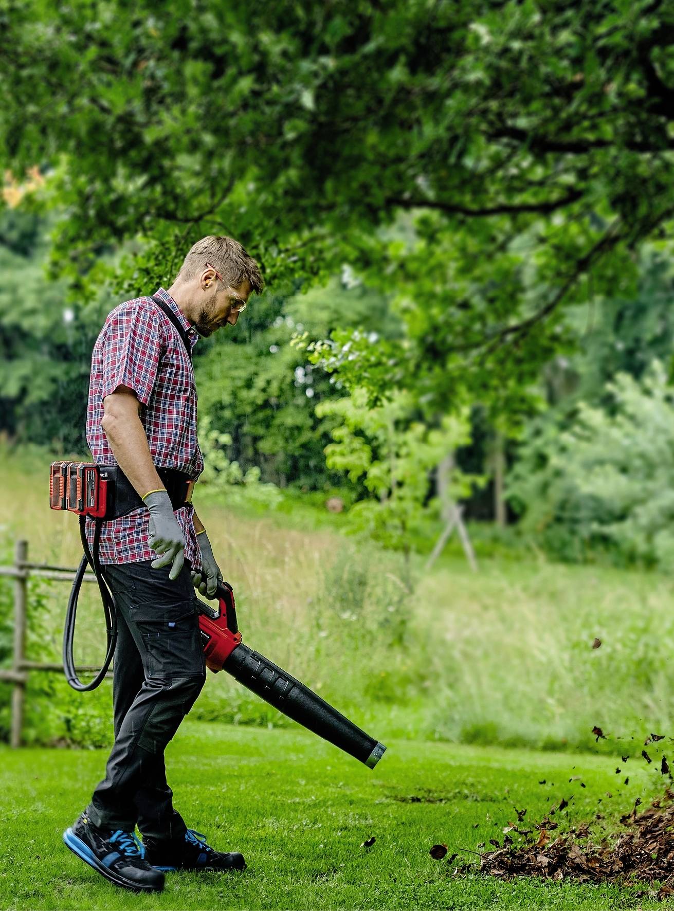 Un homme dans le jardin souffle des feuilles avec un souffleur. Il porte des gants de jardinage et a l'air concentré. L'arrière-plan est vert et boisé.