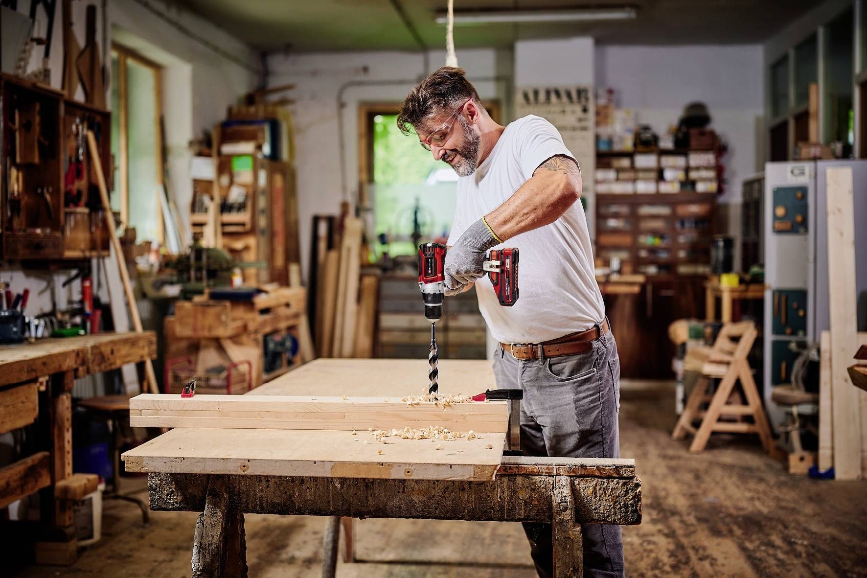 Un homme dans un atelier utilise une perceuse pour travailler une planche de bois. Entouré d'outils et de bois.