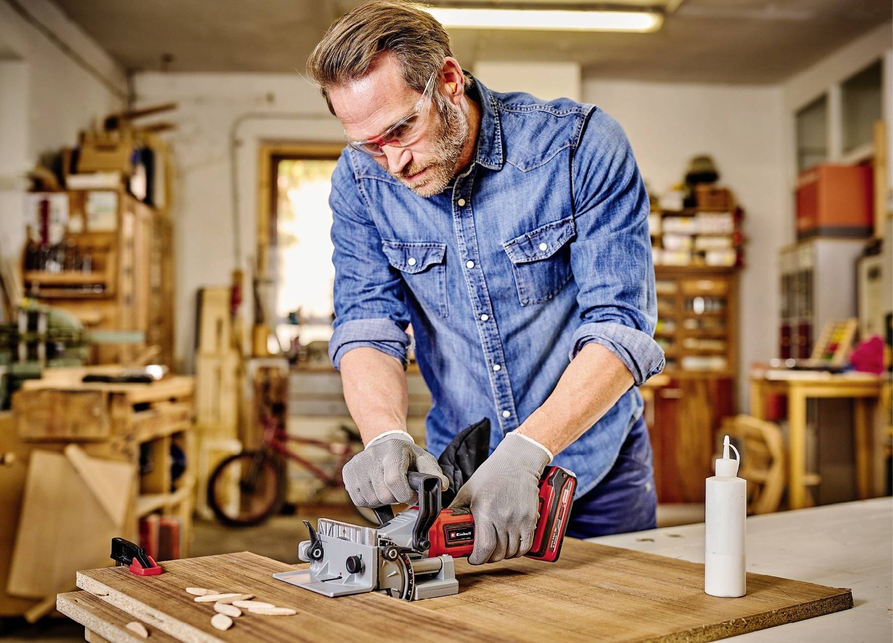 Un homme portant une veste en jean bleue et des lunettes de protection travaille une pièce de bois dans un atelier. Il utilise un outil électrique à main.