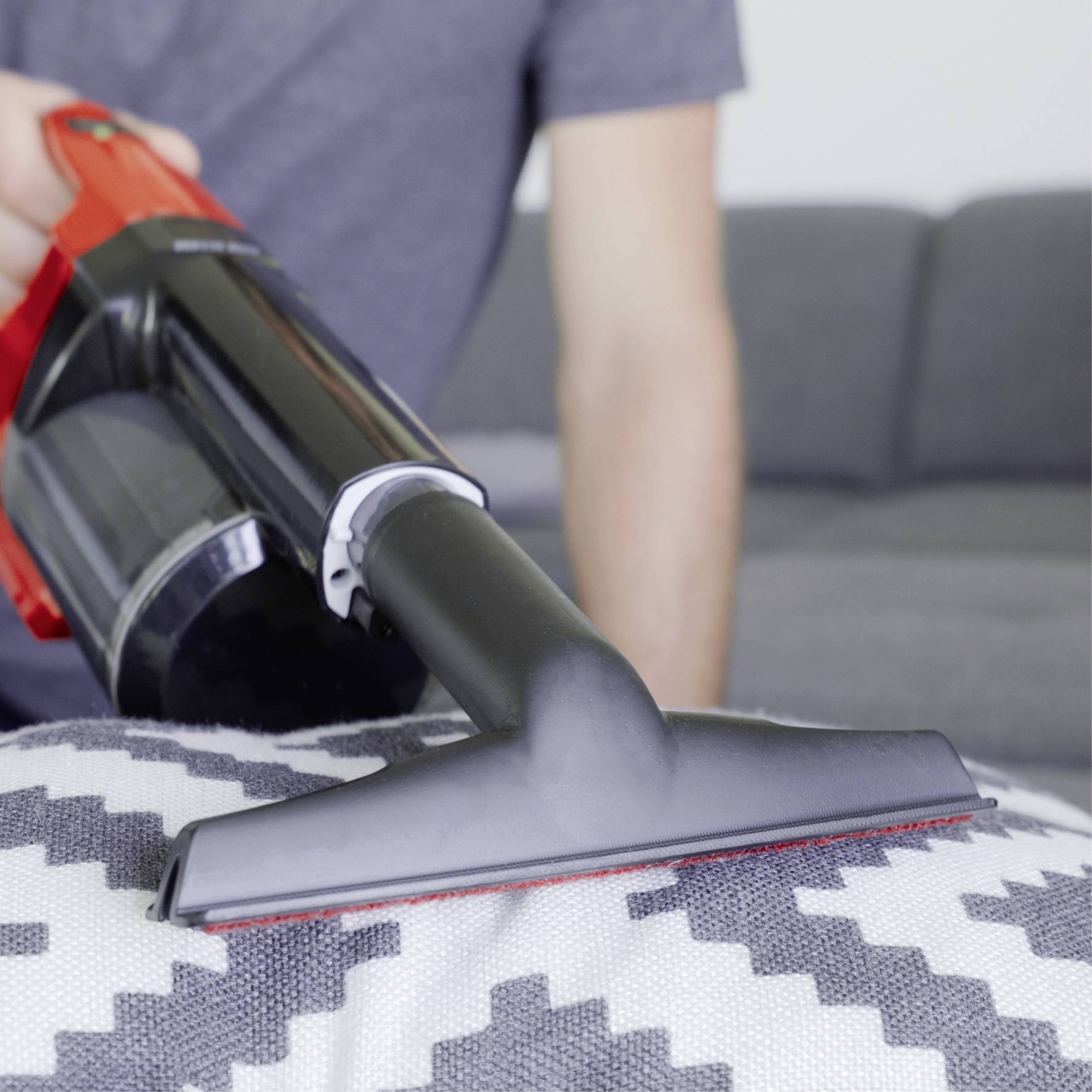 Une personne nettoie un coussin de canapé avec un aspirateur. Un canapé gris est visible en arrière-plan.