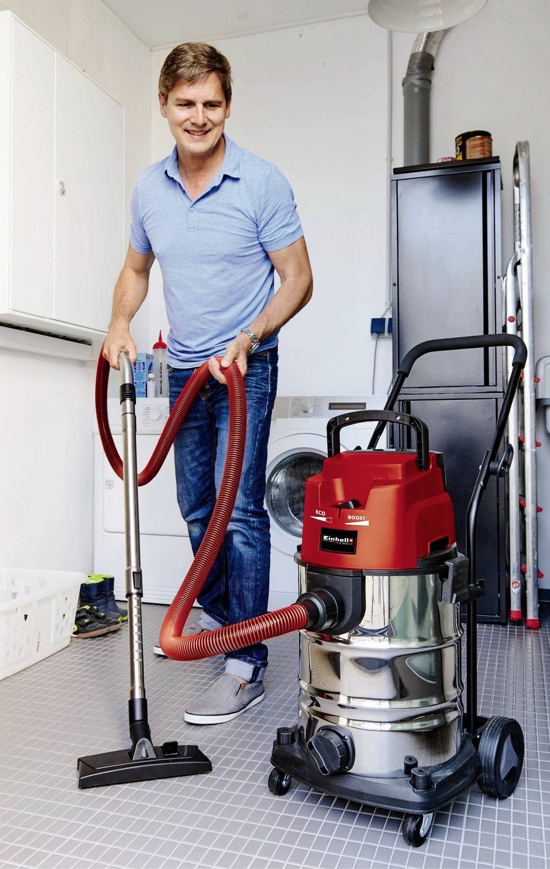 Un homme sourit en utilisant un aspirateur industriel dans une arrière-cuisine. Des appareils électroménagers sont visibles en arrière-plan.