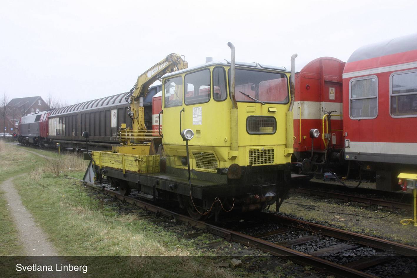 Un véhicule de service ferroviaire jaune est stationné sur une voie ferrée entre des trains rouges et noirs. Il est équipé d'un petit bras de grue. Des maisons sont visibles en arrière-plan.