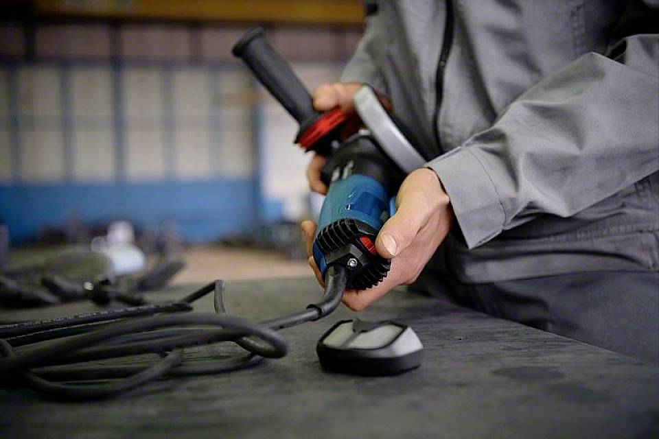 Une personne en vêtements de travail tient un outil électrique bleu avec un câble, probablement dans un environnement industriel.