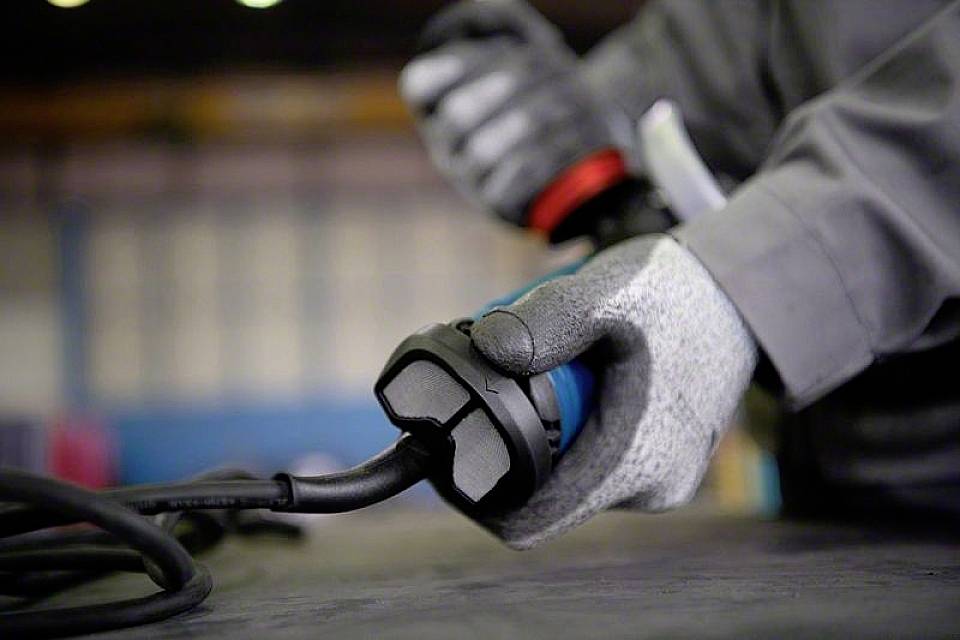 Une personne portant des gants gris tient une fiche électrique. L'arrière-plan est flou et montre un environnement industriel.