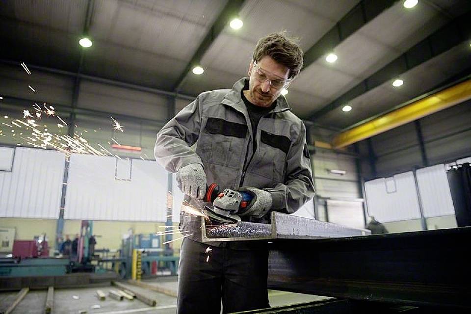 Un homme en tenue de travail coupe du métal avec une meuleuse d'angle, des étincelles volent. Il travaille dans un grand atelier industriel.
