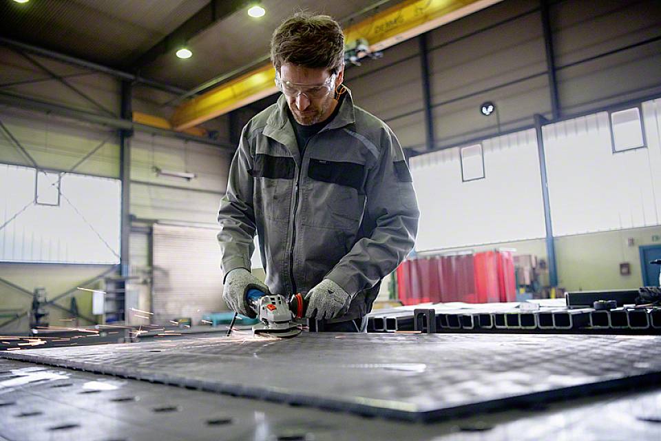 Une personne en tenue de protection meule du métal dans un hall d'usine. Des étincelles jaillissent de la meuleuse, et le hall est bien éclairé.