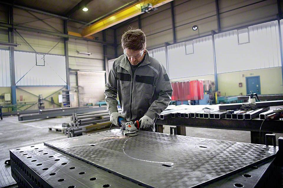 Un homme en tenue de travail usine une plaque métallique à l'aide d'un outil de meulage dans un grand atelier industriel.