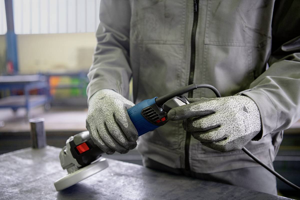 Une personne portant des vêtements de travail et des gants tient et utilise un outil électrique pour le travail des métaux dans un environnement d'atelier.