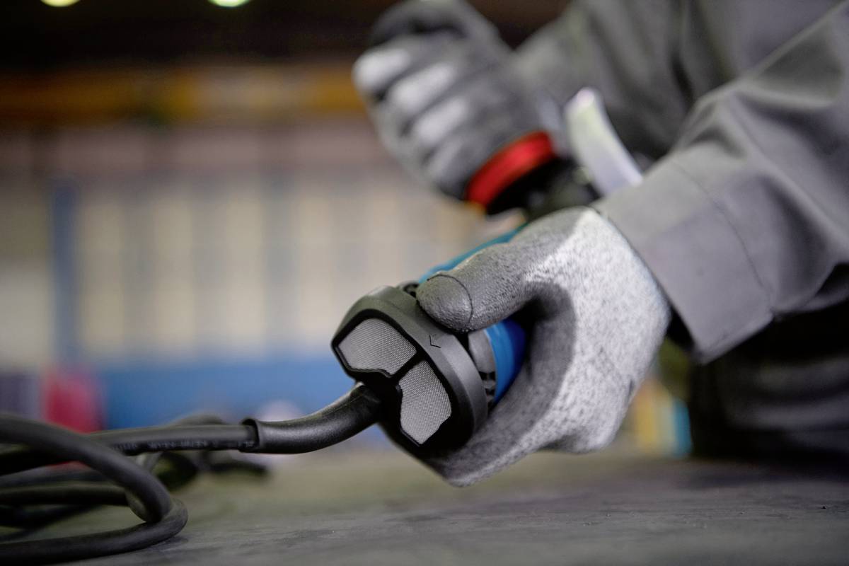 Une personne portant des gants gris tient la fiche d'un câble électrique, probablement dans un environnement d'atelier.