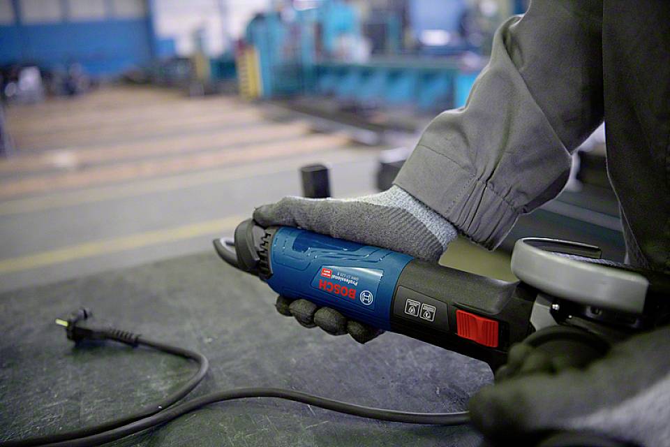 Une personne tient une meuleuse d'angle bleue dans un atelier. Un câble électrique est posé de manière lâche sur la table devant elle.