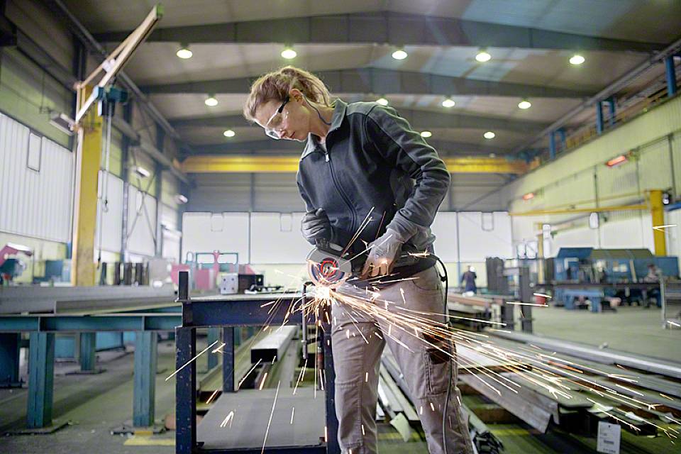Une personne travaille avec une meuleuse d'angle dans un grand atelier bien éclairé ; des étincelles volent pendant que le métal est découpé.