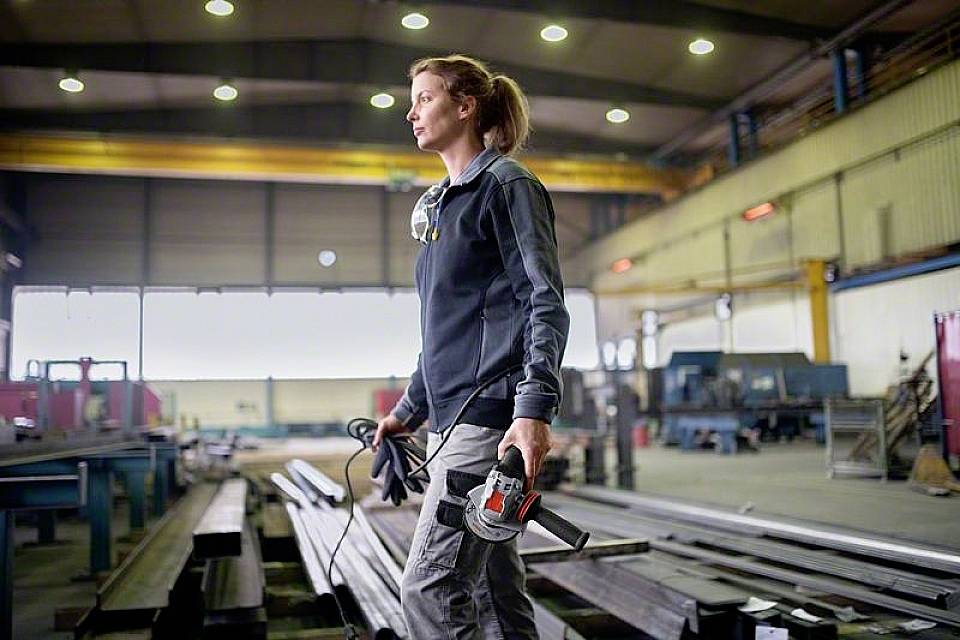 Une femme en tenue de travail se tient dans une halle industrielle et tient un outil électrique. La halle est vaste et a un aspect industriel.
