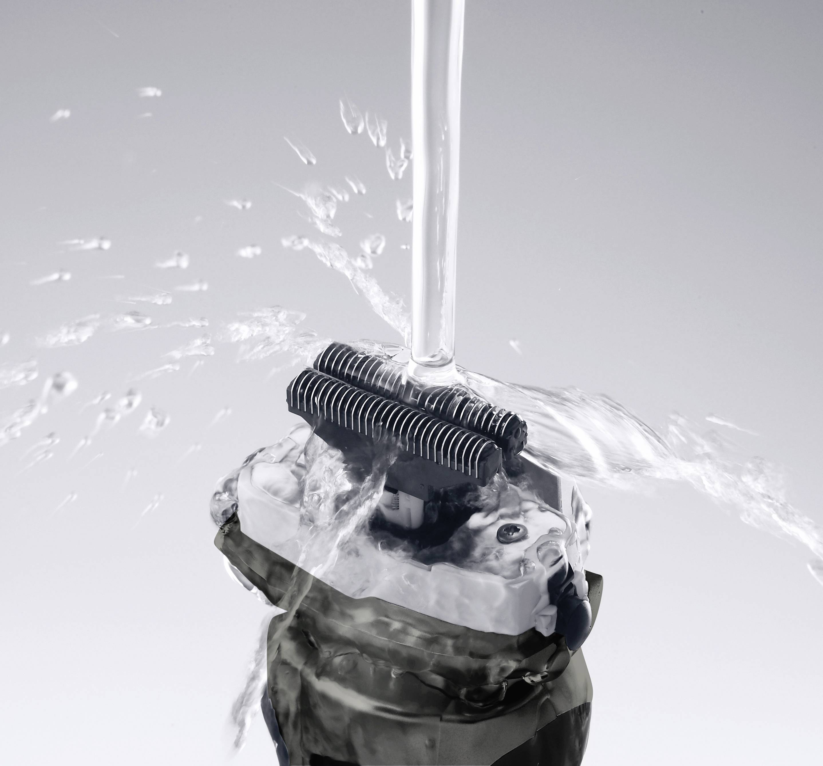 Rasoir électrique nettoyé sous un jet d'eau. L'eau gicle autour des lames qui sont rincées pour assurer leur propreté.